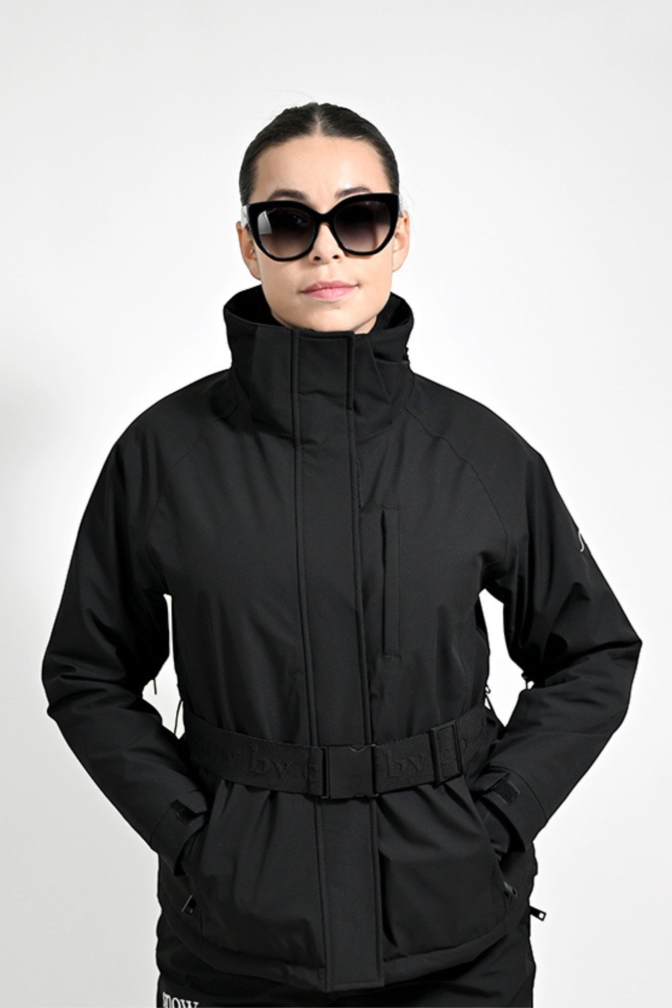 Snow by Clo black waterproof ski jacket with a belted waist and high collar worn by a woman in sunglasses.